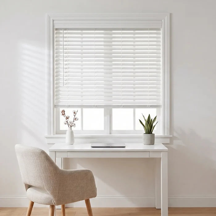 White window blinds installed on a white-framed window in a bright, modern room with a white desk, beige upholstered chair, and small potted plant.