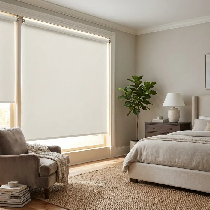 A bright, airy bedroom featuring milky white Luxe fabric blackout roller shades, styled with a neutral armchair and a large indoor plant
