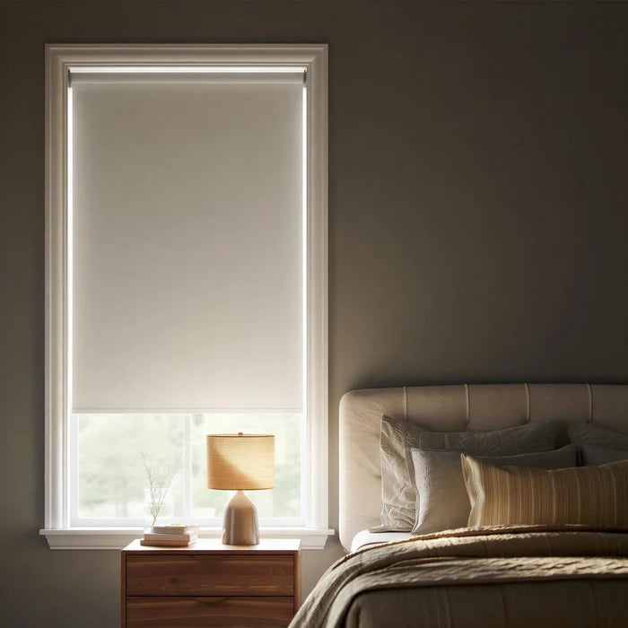 Off-center angled shot of a white elegance room darkening roller shade in a modern bedroom with a bed and pillows visible on the right